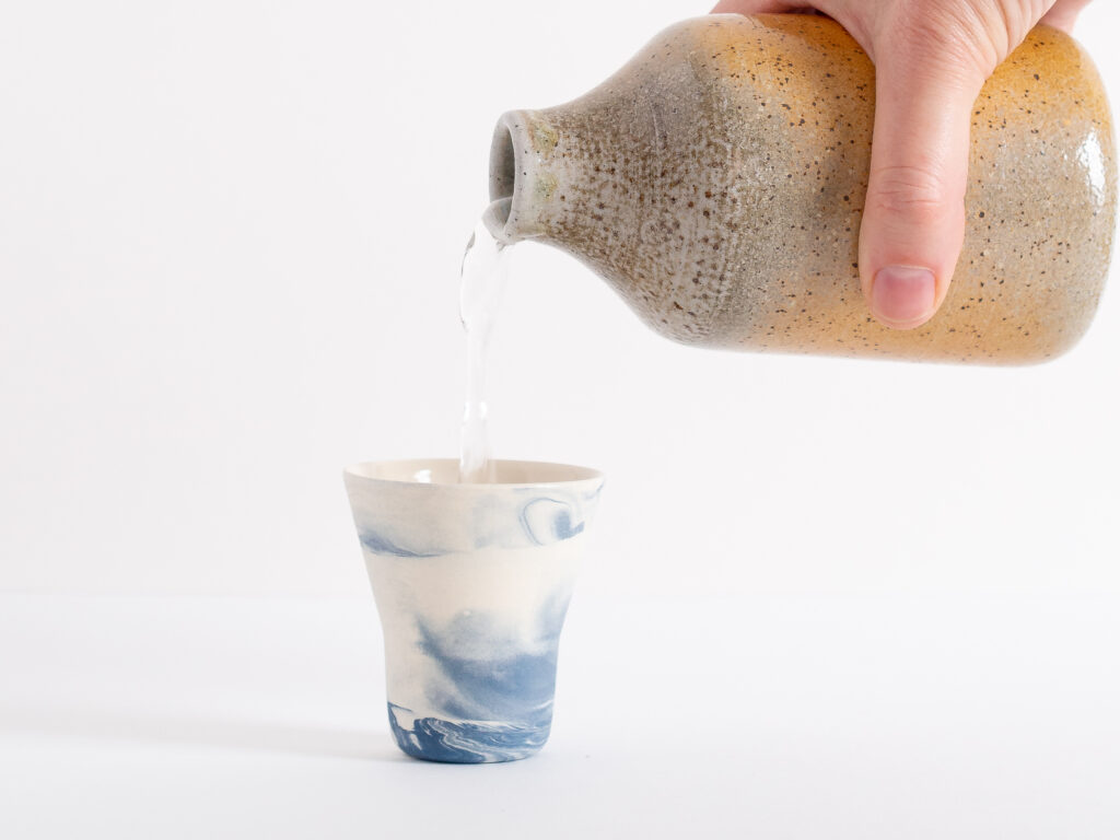 A sake cup with marbled blue patter and a hand holding a ceramic bottle pouring sake into the cup